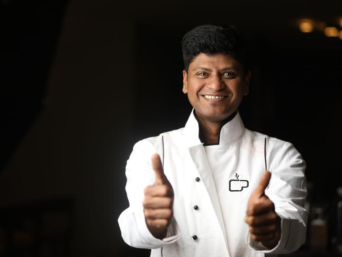 Smiling Indian chef in a white chef's coat giving a thumbs up in a dark kitchen