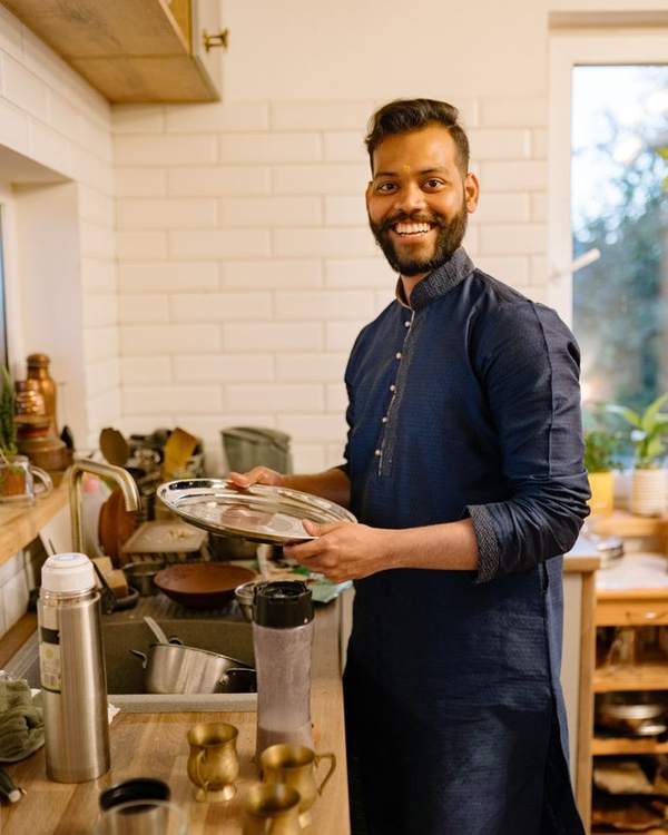 Smiling Indian chef in a navy apron holding a tray in a bright kitchen