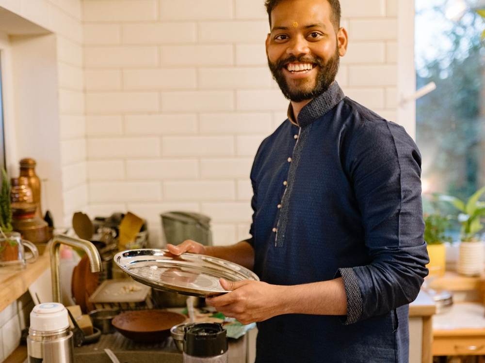 Smiling Indian chef in a dark apron holding a tray of dishes in a home-style kitchen