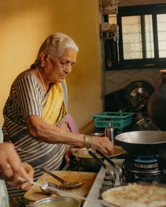 An older Indian woman in a yellow saree cooking at a stove in a home kitchen