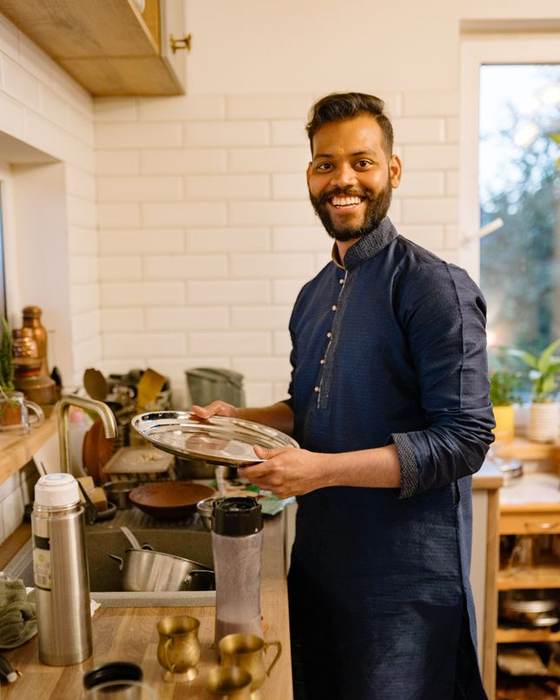 Smiling Indian chef in a dark apron holding a tray of dishes in a home-style kitchen