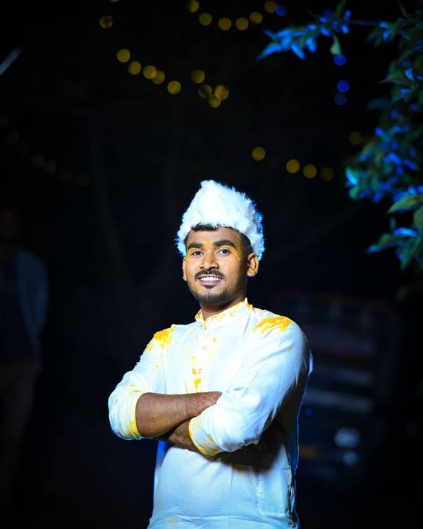 Indian man in a white kurta and cap standing with arms crossed under fairy lights