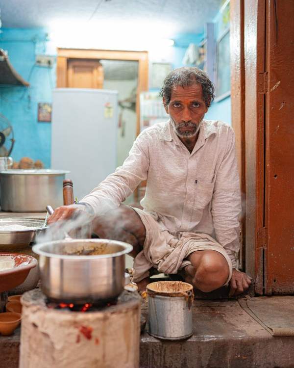 Elderly Indian cook in a pink shirt working over steel pots in a small kitchen