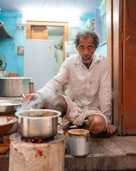 Indian cook seated with large steel pots in a blue-walled kitchen