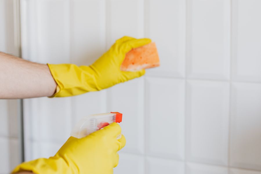 Yellow-gloved hands cleaning white bathroom tiles with a sponge and spray