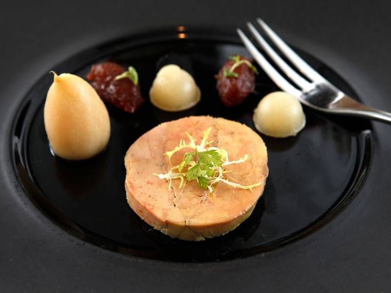 A tasting course plated on a dark plate with tweezers placing a garnish