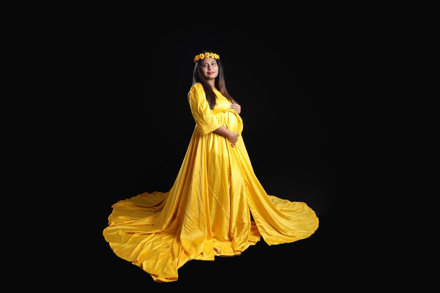 A studio maternity portrait of a woman in a flowing yellow gown against a black background