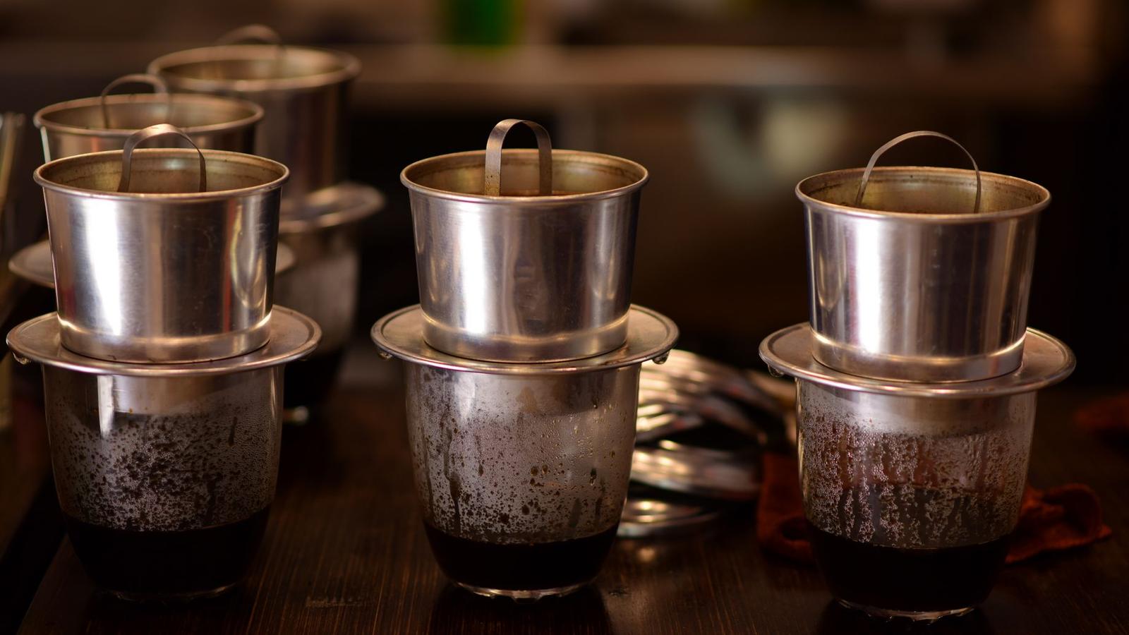 Three South Indian steel filter coffee sets on a dark wooden counter