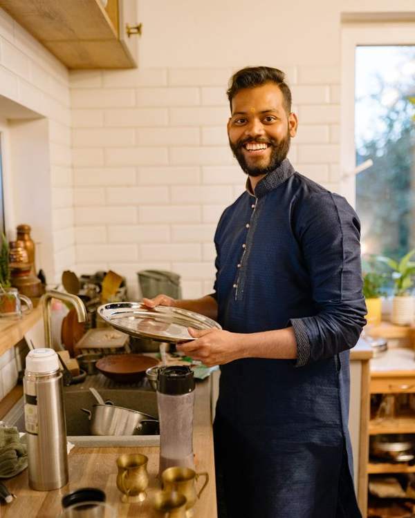 Smiling Indian chef in a navy apron holding a pan in a bright kitchen