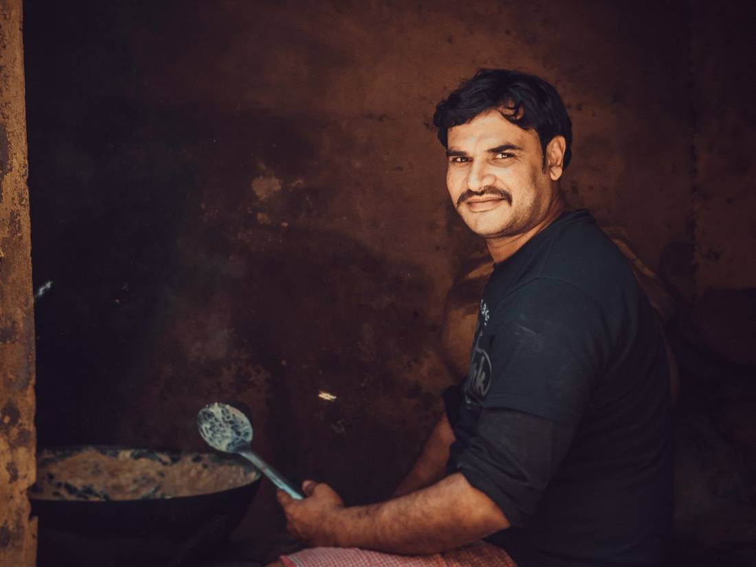 Indian chef seated by a wood fire holding a ladle, lit warmly from the flames