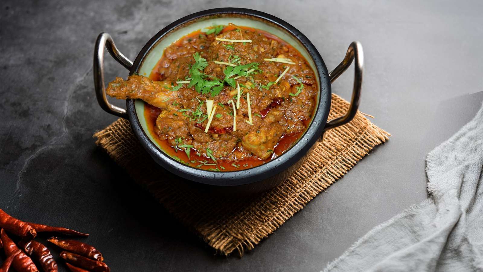 Chettinad chicken curry in a black iron kadhai with dried red chillies on a grey stone table