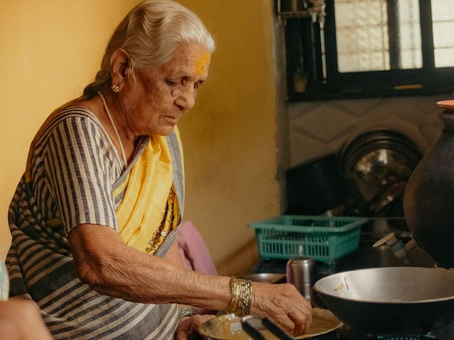 Elderly Indian woman cooking at a stove in a sunlit yellow kitchen