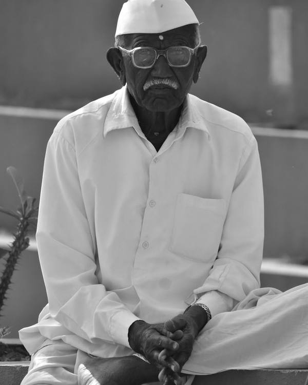 Elderly Indian man in a white shirt and cap, seated, in black and white