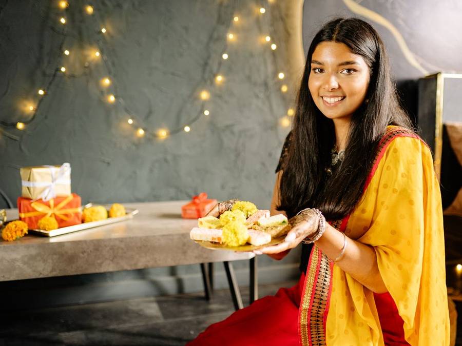 Indian woman in a yellow kurta holding a platter of sweets beside fairy lights