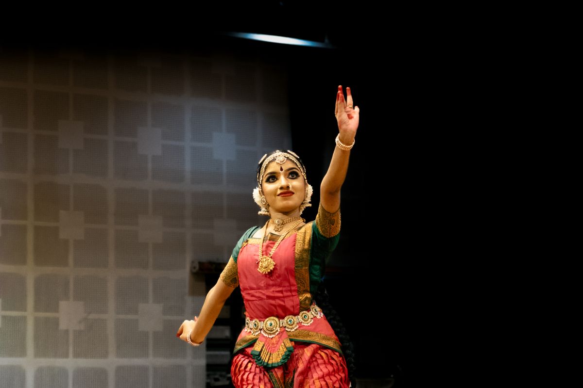 A Bharatanatyam dancer in a rose-and-gold costume performing on a dim stage