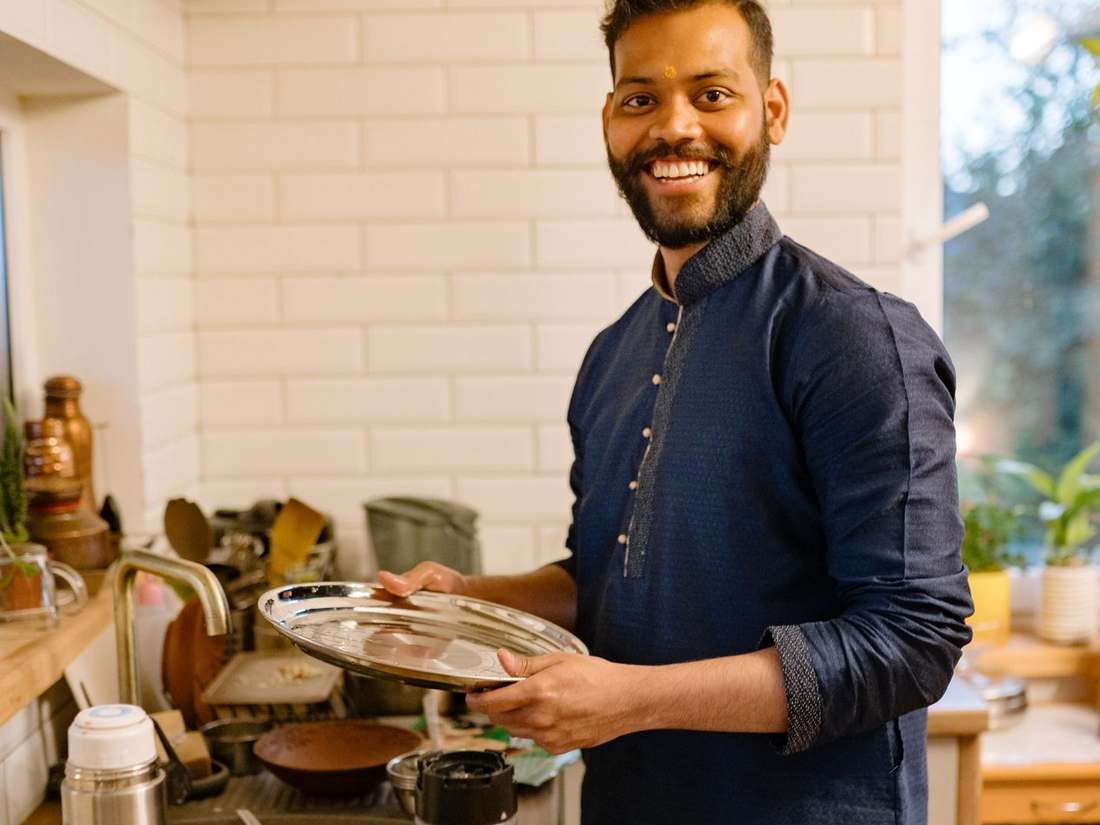 Smiling Indian chef in a blue shirt holding a plate in a bright kitchen