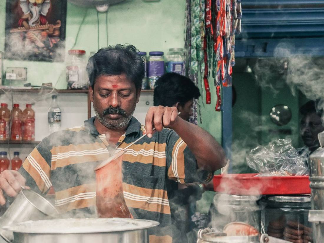 Indian chaiwala straining chai at his stall, steam rising from the pan